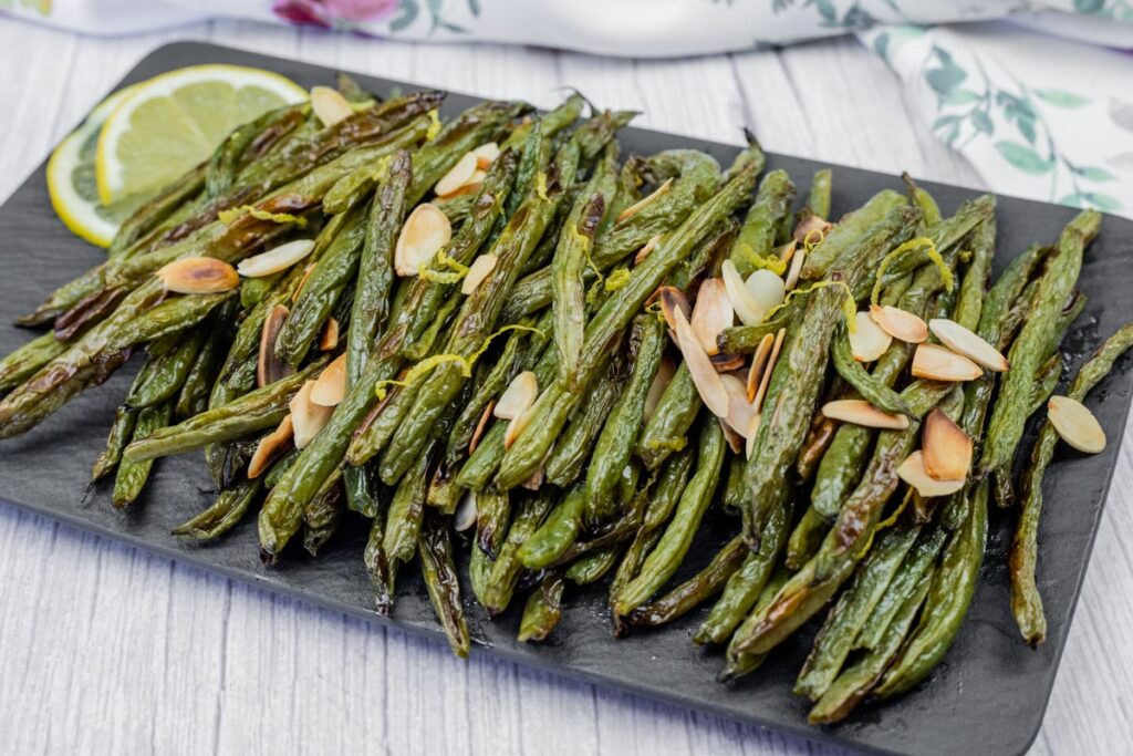 A platter of roasted green beans topped with sliced almonds, garnished with a lemon slice, on a dark rectangular serving tray.