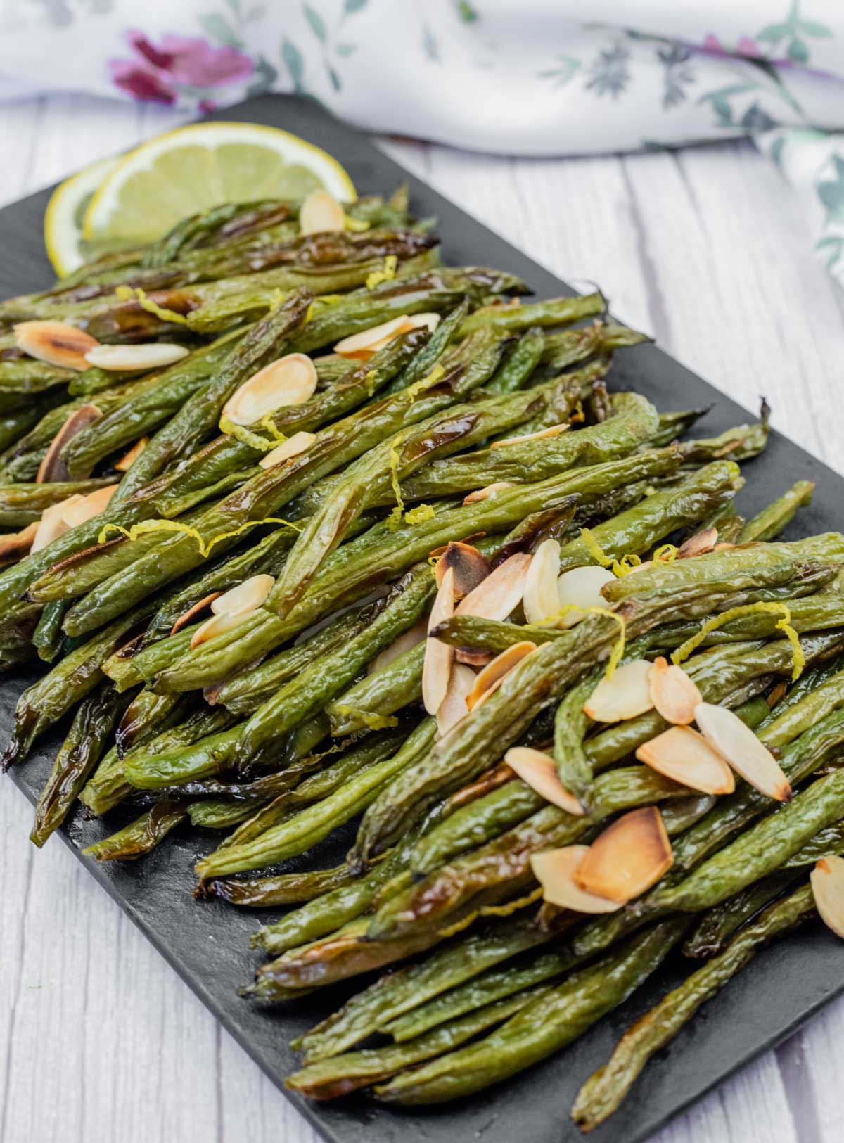 A platter of roasted green beans topped with sliced almonds and lemon zest. A lemon slice is in the background.