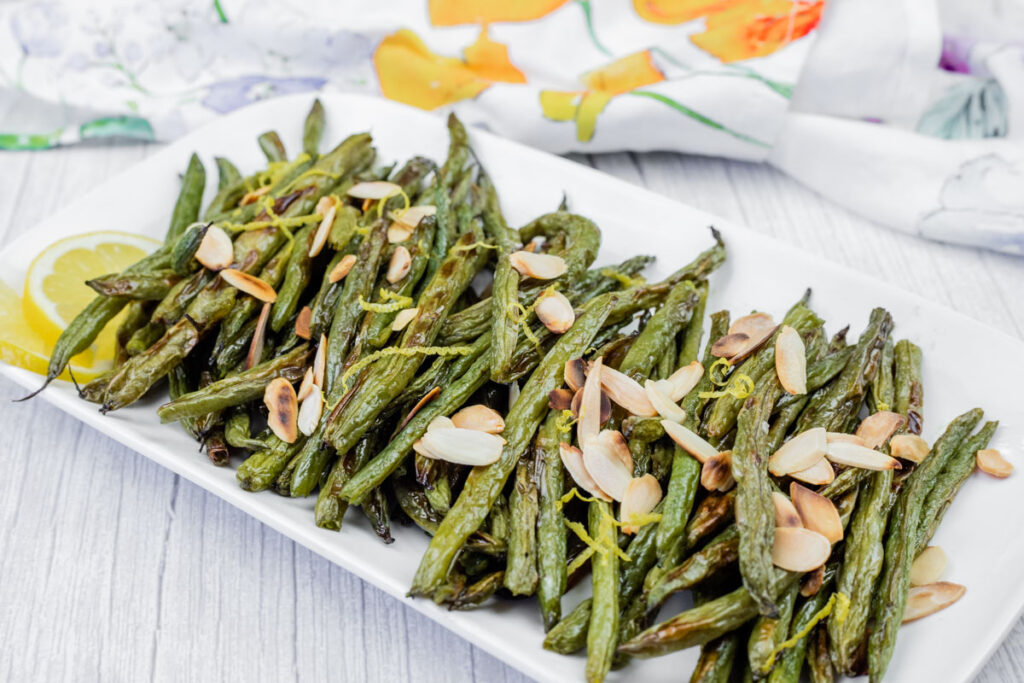 Roasted green beans with sliced almonds and lemon wedges on a rectangular white plate, placed on a light wooden surface.