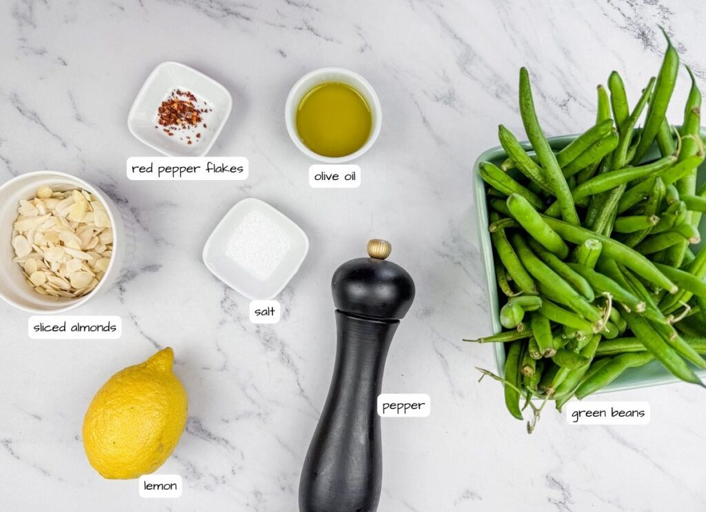 Ingredients on a marble surface: red pepper flakes, olive oil, green beans, sliced almonds, salt, lemon, and a pepper grinder.