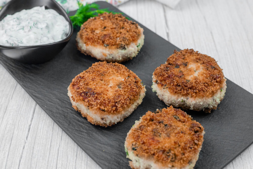 Four Salmon Cakes are arranged on a rectangular black plate next to a small bowl of creamy, herb-speckled sauce. The patties appear golden brown and crispy.