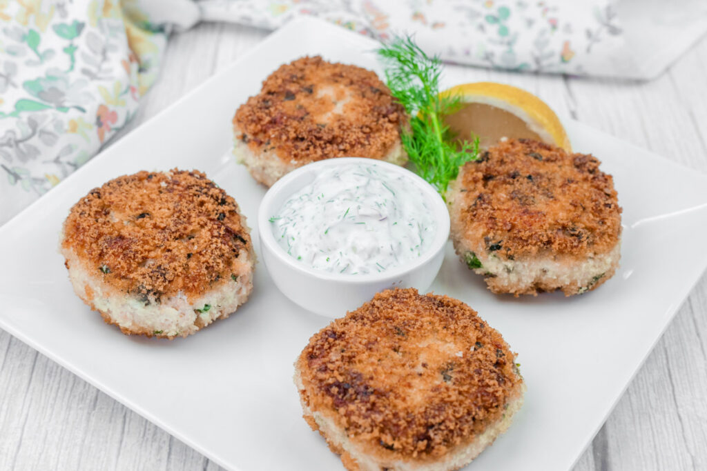 Five Salmon Cakes on a square white plate with a small bowl of creamy sauce, garnished with a lemon wedge and dill.