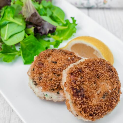 Two Salmon Cakes on a white plate with mixed greens and a lemon wedge.