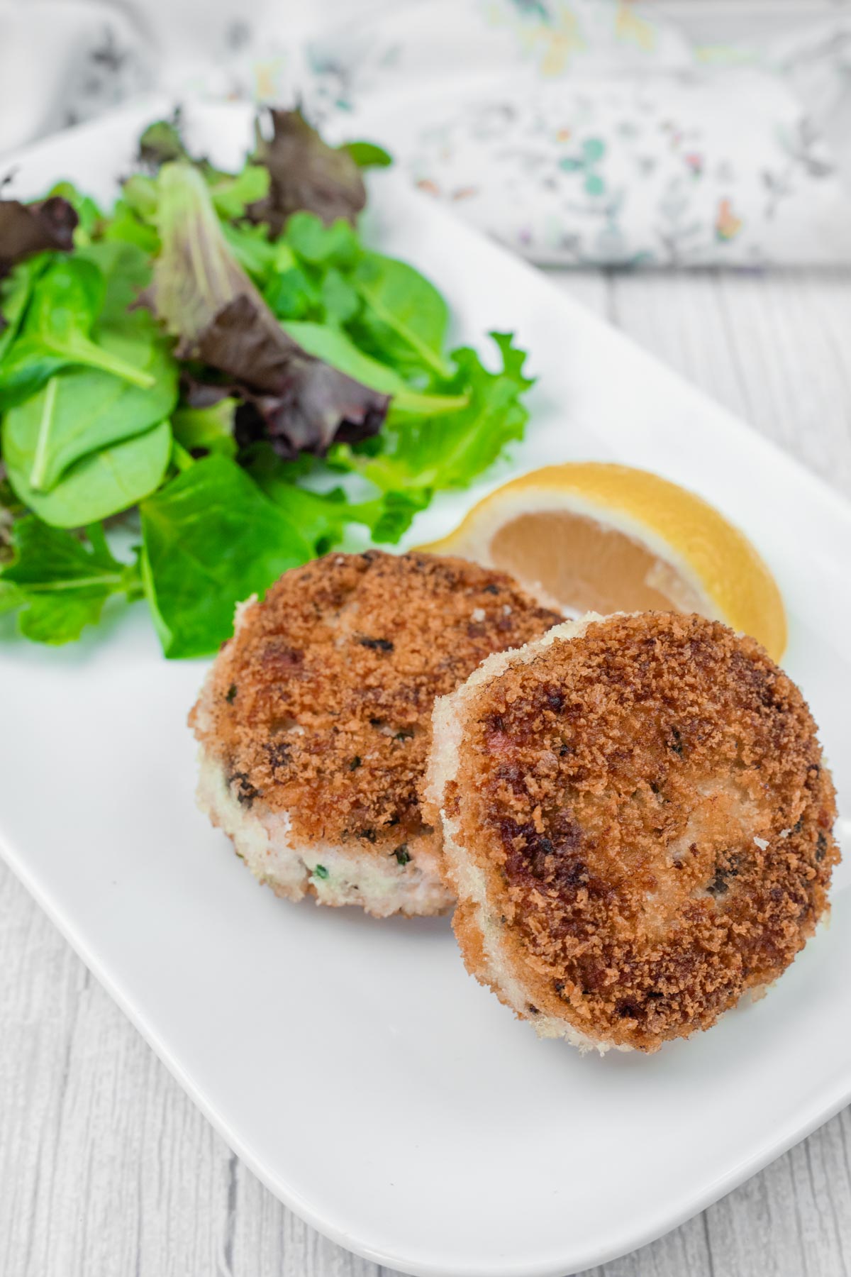 Two Salmon Cakes on a white plate with mixed greens and a lemon wedge.