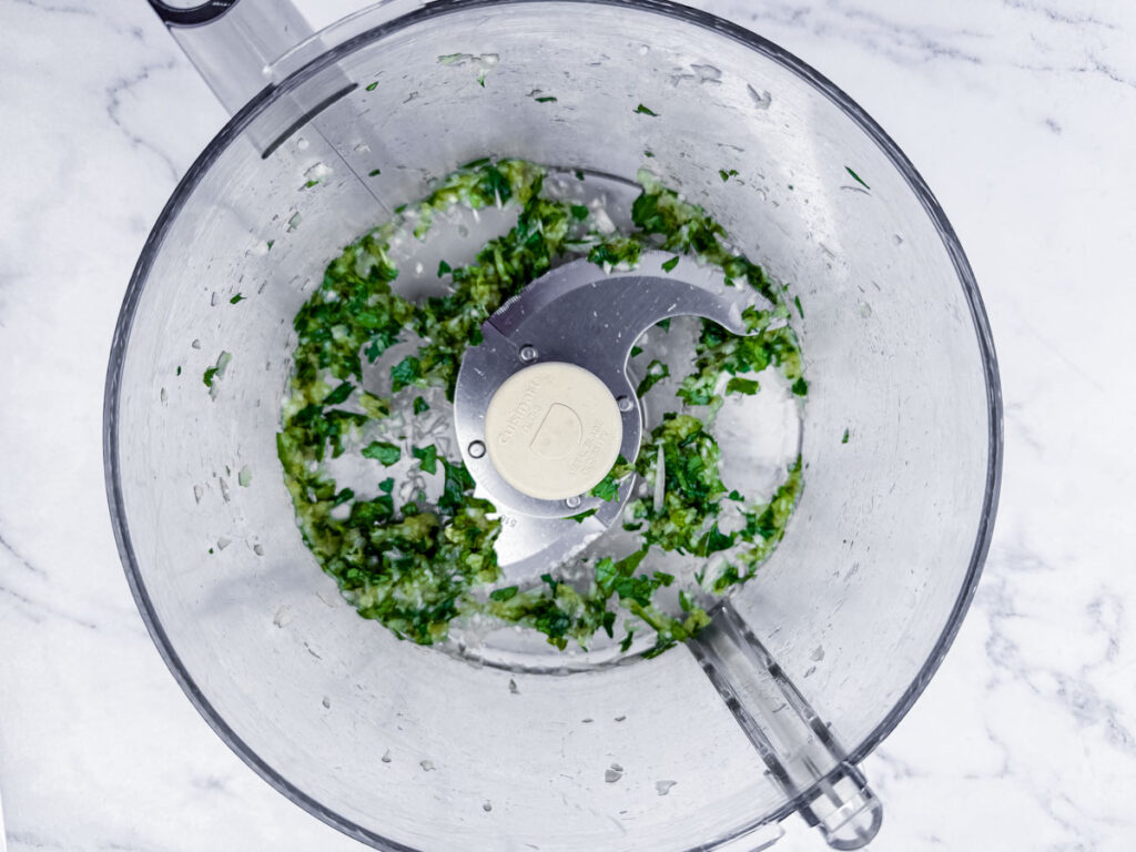 Top view of a food processor containing chopped green herbs, likely parsley or cilantro, on a white marble surface.