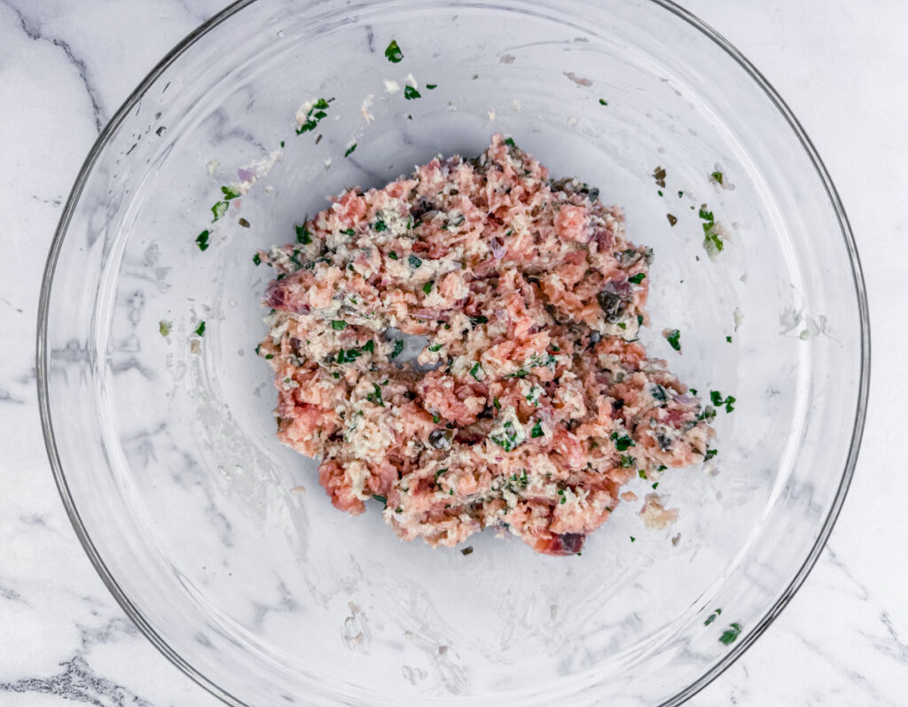 A glass bowl contains a mixture of minced meat, herbs, and spices on a marble surface.