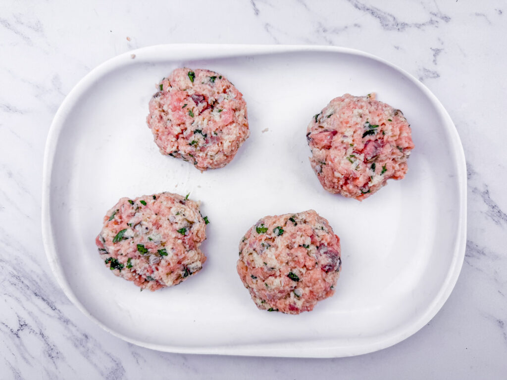 Four raw meat patties mixed with herbs, arranged on a white oval plate. The background is a marble surface.