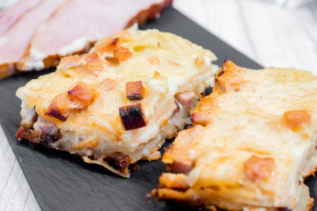 Close-up of two slices of Scalloped Potatoes with Ham and Cheese on a black plate, with sliced ham in the background.