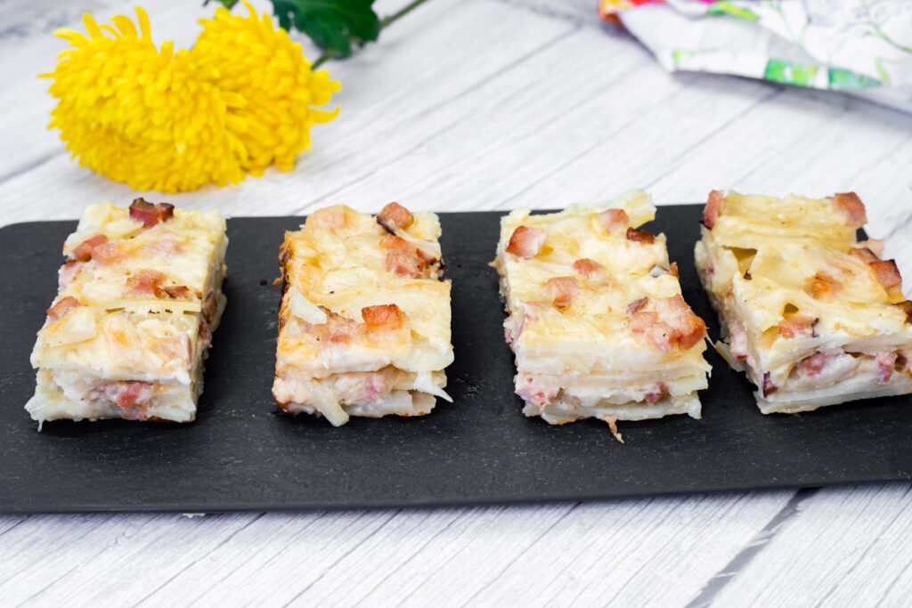 Four slices of Scalloped Potatoes with Ham and Cheese on a black slate board, placed on a light wooden surface. Bright yellow flower in the background.