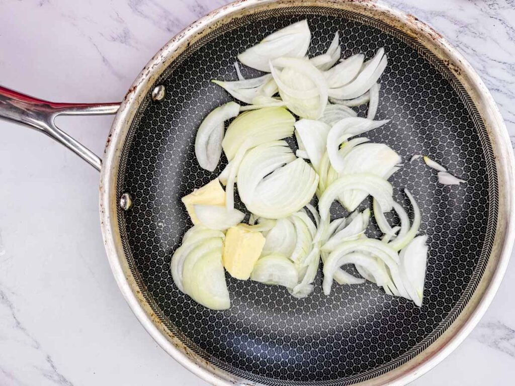 Sliced onions and butter in a frying pan on a marble counter.