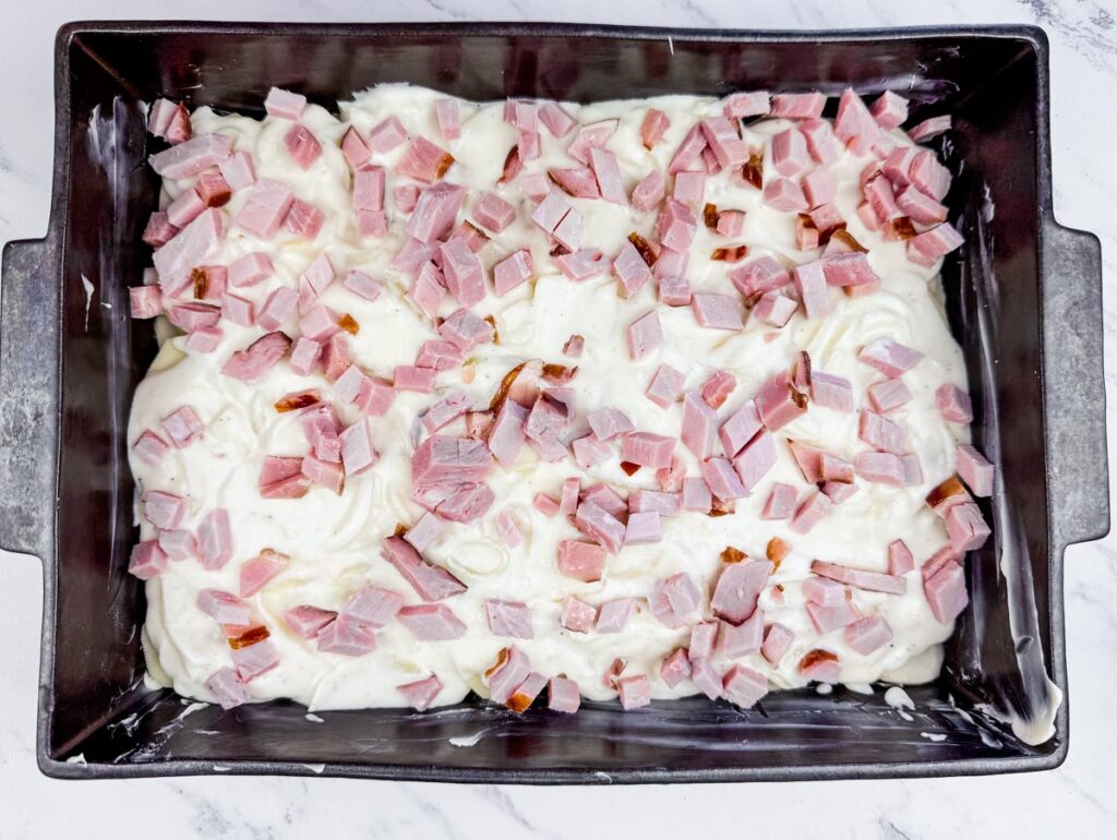 A baking dish with diced ham pieces on top of a creamy white mixture, placed on a marble surface.