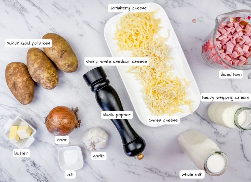 Ingredients for a recipe arranged on a marble surface: Yukon Gold potatoes, black pepper, diced ham, heavy whipping cream, whole milk, cheeses, butter, onion, garlic, and salt.