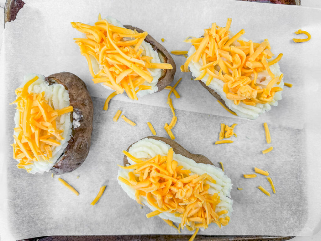 Four baked potatoes topped with mashed potatoes and shredded cheddar cheese on parchment paper.