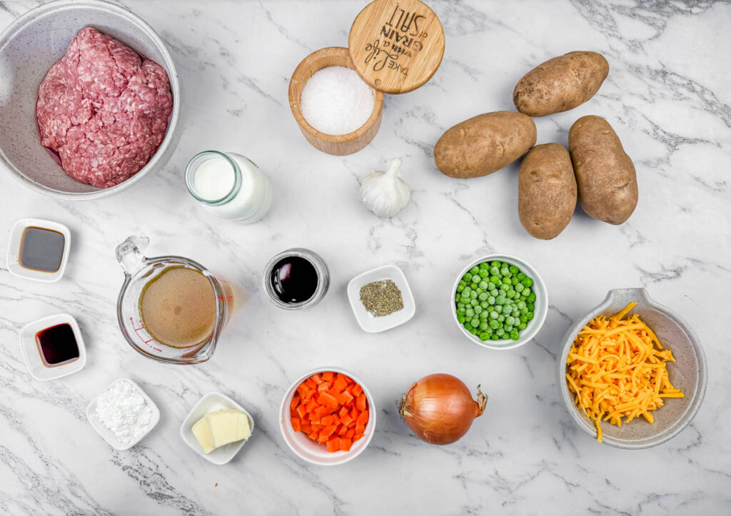 Ingredients for Shepherd’s Pie Baked Potatoes arranged on a marble surface, including ground meat, potatoes, onion, carrots, peas, butter, broth, cream, soy sauce, garlic, salt, pepper, cornstarch, and cheese.