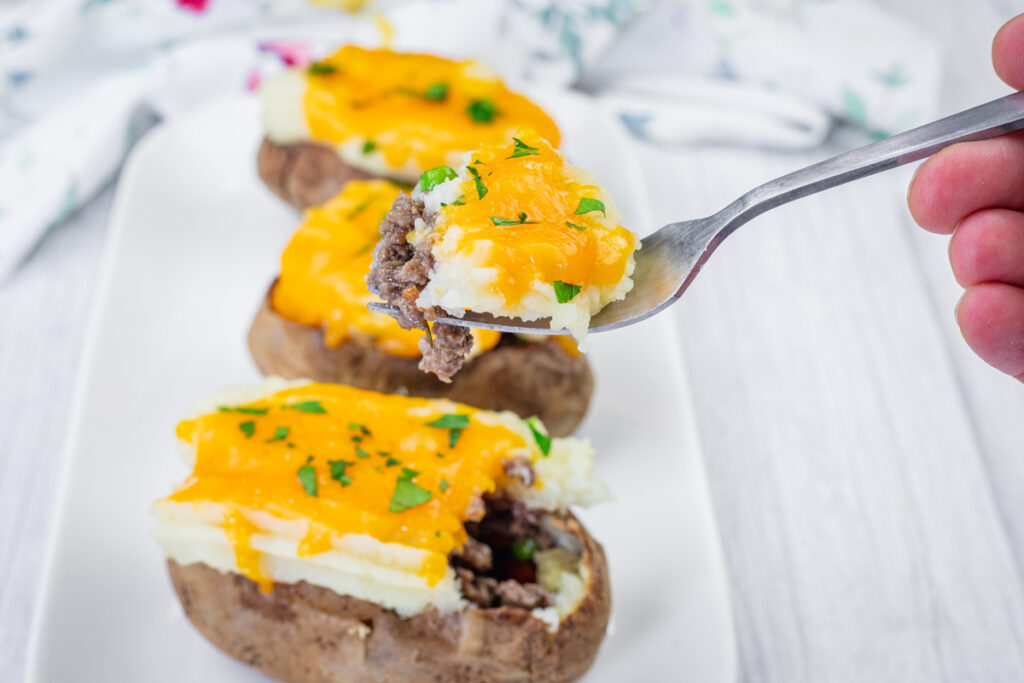 A close-up of Shepherd’s Pie Baked Potatoes topped with ground beef, melted cheddar cheese, and chopped parsley on a white plate. A fork holds a piece with all toppings.