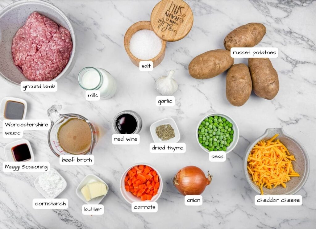 Ingredients for Shepherd’s Pie Baked Potatoes displayed on a marble surface, including ground lamb, potatoes, peas, carrots, onion, garlic, cheddar cheese, milk, butter, red wine, broth, seasonings, and salt.