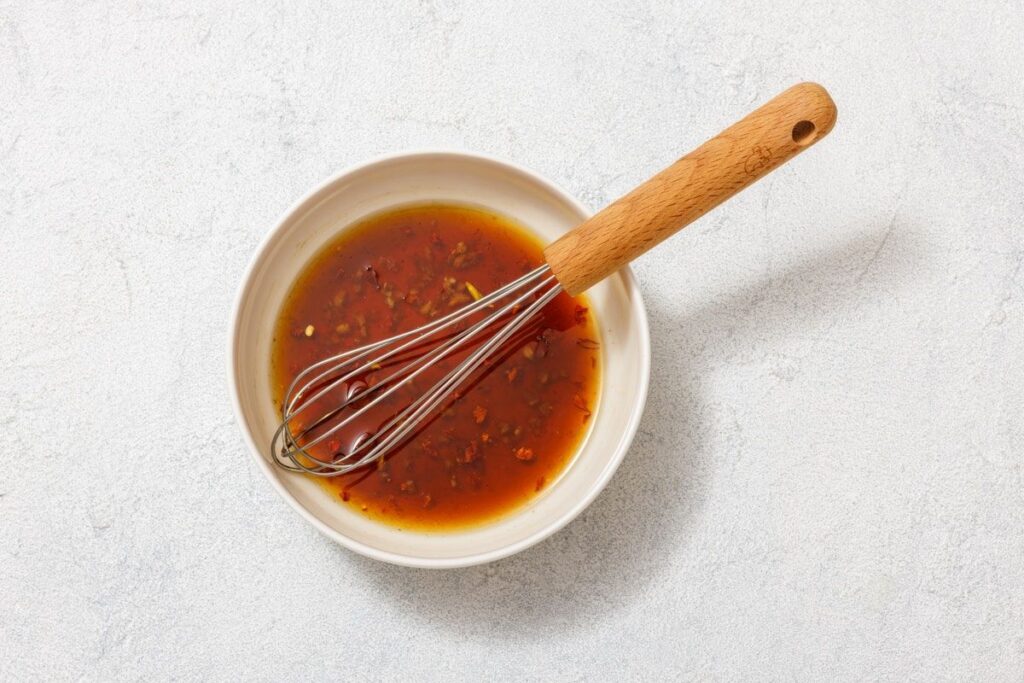 A white bowl with brown liquid marinade and visible herbs, containing a whisk with a wooden handle, sits on a light surface.