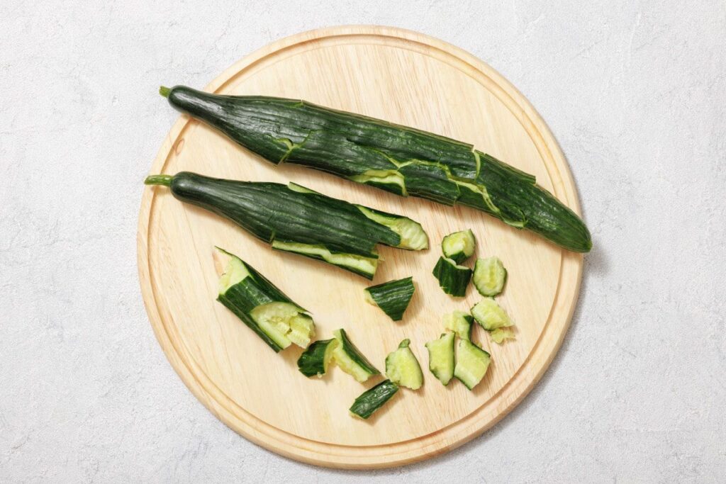 Two partially peeled cucumbers and sliced pieces are on a round wooden cutting board against a light gray background.