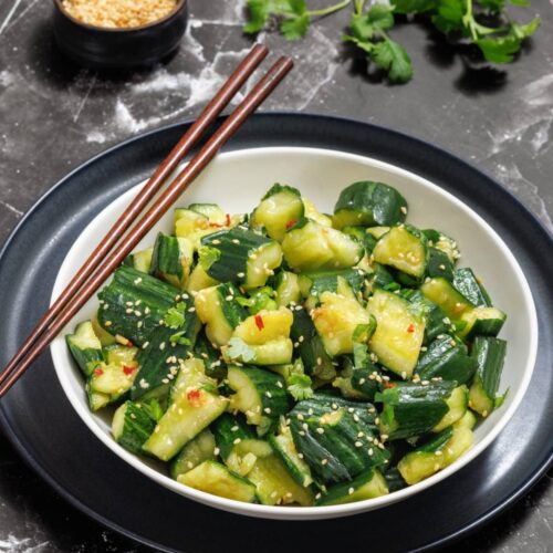 A bowl of smashed cucumber salad with sesame seeds on a dark, marbled surface. Chopsticks rest on the bowl's rim. A small bowl of sesame seeds and cilantro leaves are in the background.