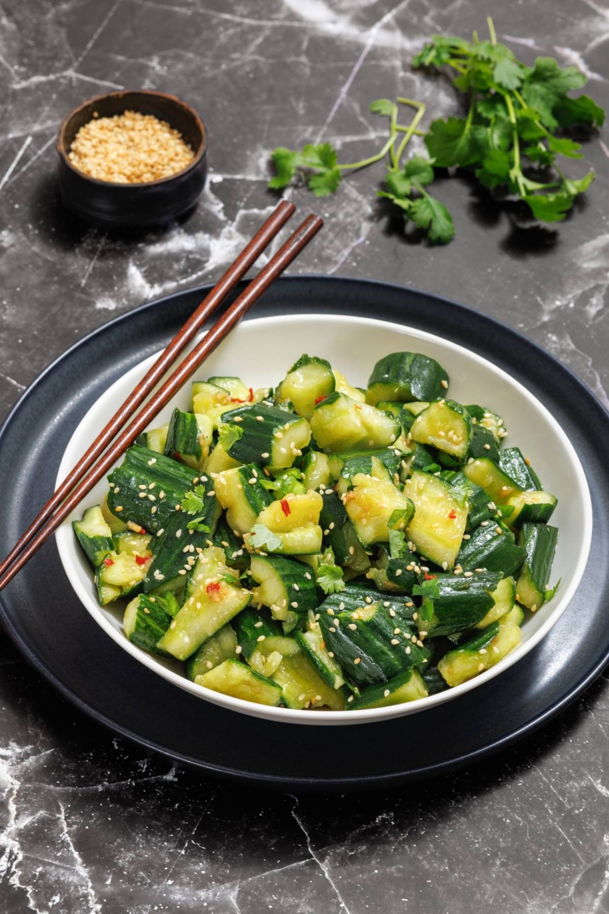 A bowl of smashed cucumber salad with sesame seeds on a dark, marbled surface. Chopsticks rest on the bowl's rim. A small bowl of sesame seeds and cilantro leaves are in the background.