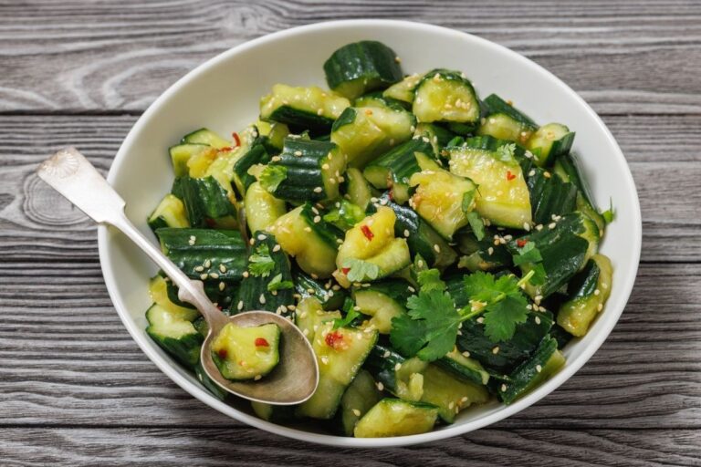 A bowl of diced cucumbers with sesame seeds, cilantro, and red pepper flakes, accompanied by a silver spoon, on a wooden surface.