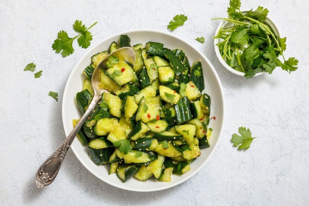 A bowl of cucumber salad with chopped cilantro and chili flakes, accompanied by a small bowl of fresh cilantro on a light gray background. Silver spoon in the salad.