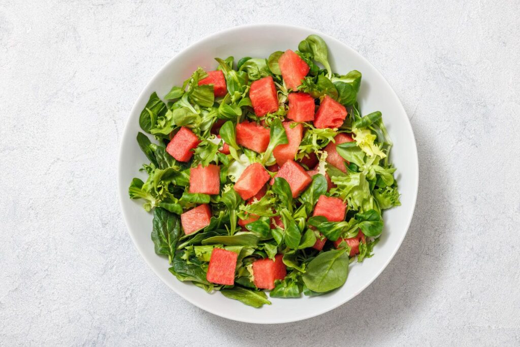 A bowl of salad with leafy greens and watermelon cubes on a white surface.