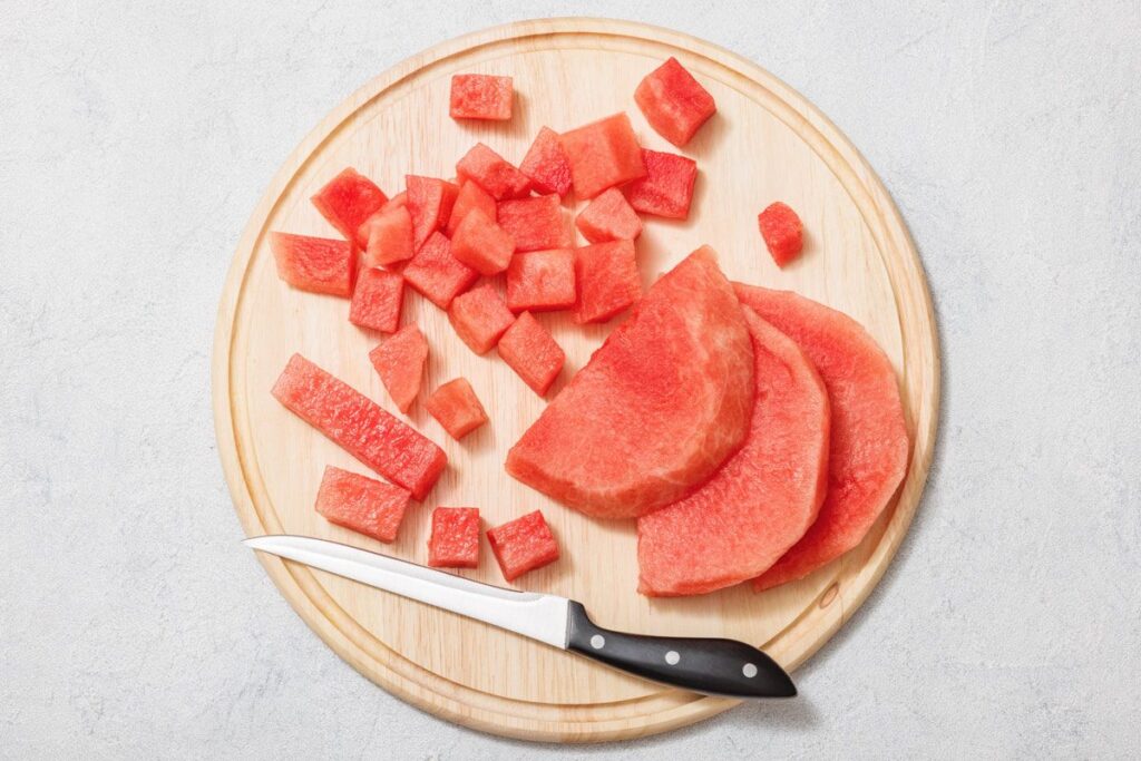 Wooden cutting board with sliced and cubed watermelon and a knife on a light background.
