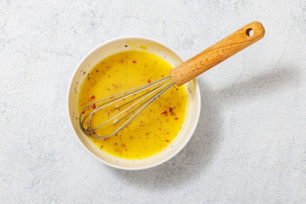 A bowl of yellow sauce mixed with herbs and spices, accompanied by a metal whisk with a wooden handle resting inside.