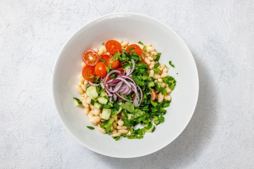 A white bowl filled with a colorful salad of white beans, cherry tomatoes, cucumber, red onion, and fresh parsley, on a light surface.