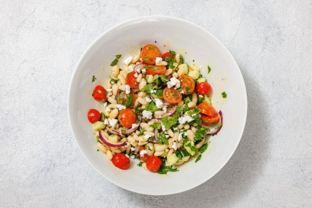 A white bowl filled with a colorful salad featuring cherry tomatoes, white beans, cucumbers, red onion, and crumbled feta cheese.