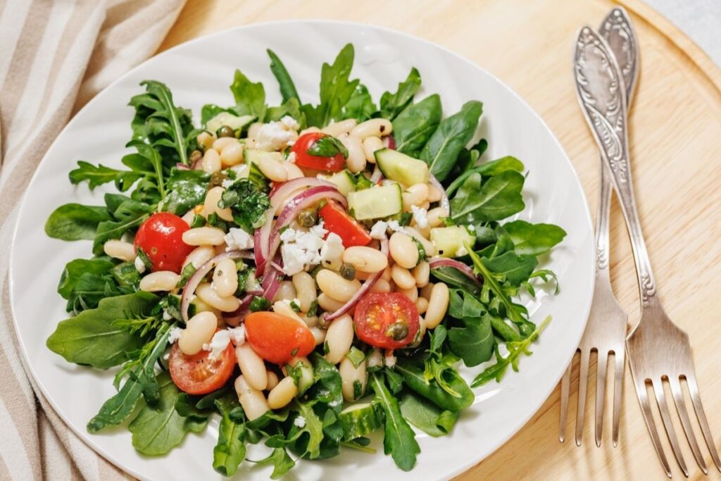 Fresh salad with arugula, cherry tomatoes, cucumbers, and white beans, garnished with red onions and feta cheese on a white plate.