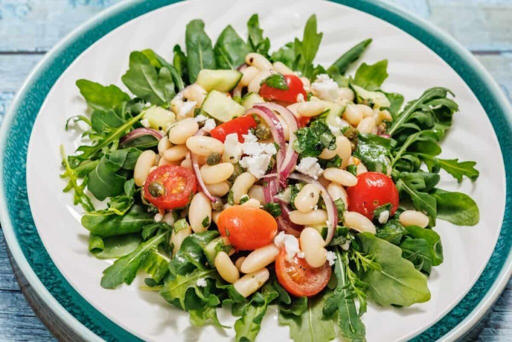 A plate of White Bean Salad with arugula, white beans, cherry tomatoes, red onion, cucumber, and crumbled cheese.