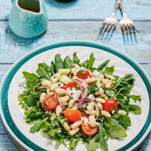 A fresh salad with arugula, cherry tomatoes, white beans, red onion, cucumber, feta cheese, and capers served on a white plate.