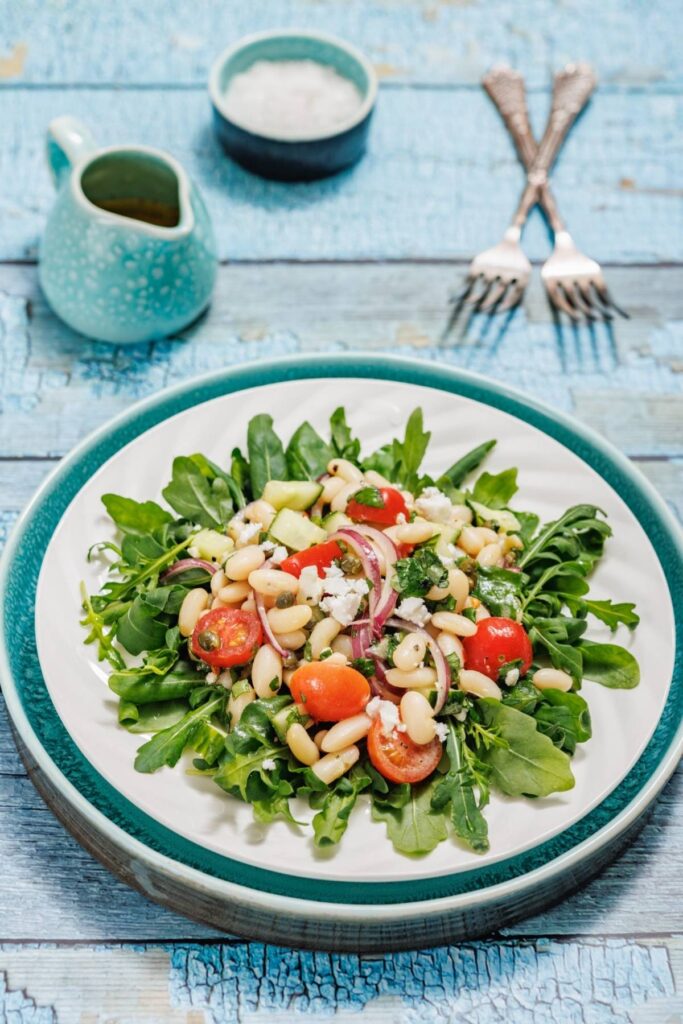A fresh salad with arugula, cherry tomatoes, white beans, red onion, cucumber, feta cheese, and capers served on a white plate.