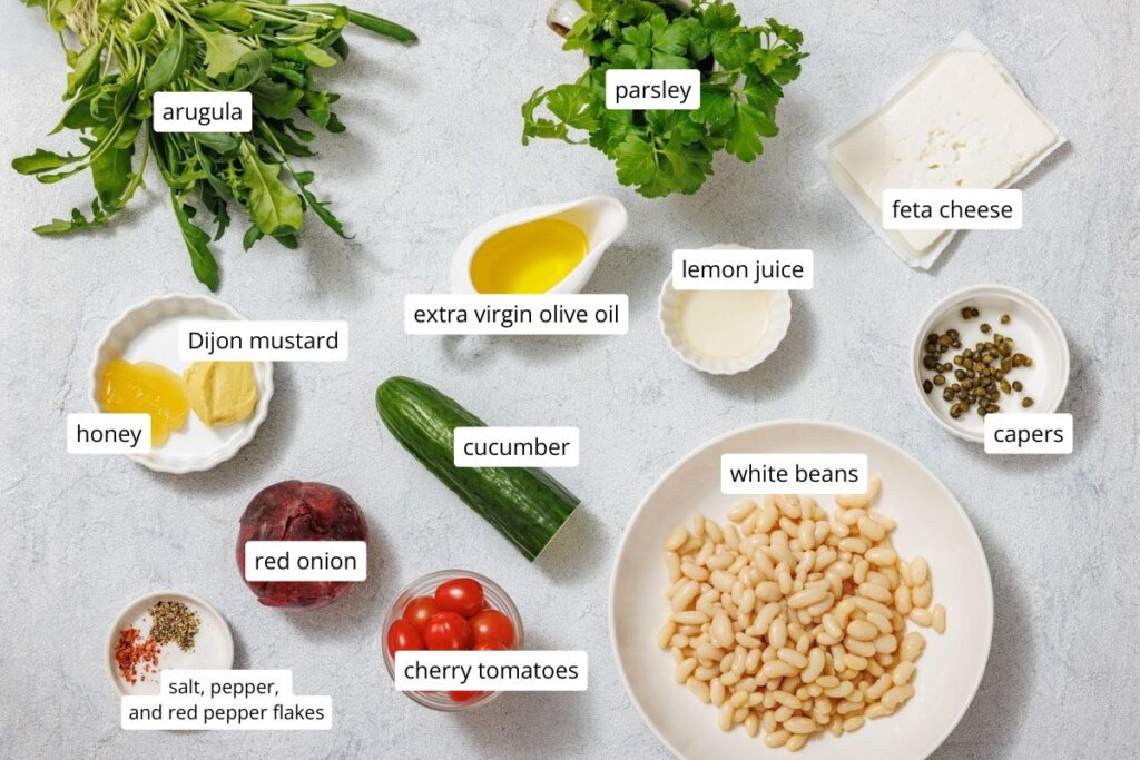 A variety of fresh ingredients for a salad, including arugula, parsley, cucumber, cherry tomatoes, white beans, and dressing items.