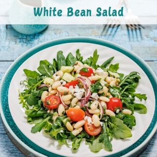 Plate of white bean salad with arugula, cherry tomatoes, red onion, and feta cheese on a blue table.