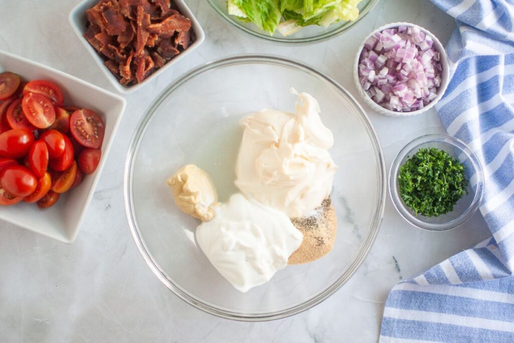 Overhead shot of a glass bowl containing creamy salad dressing ingredients, surrounded by bowls of bacon, lettuce, red onion, parsley, and tomatoes on a marble surface.
