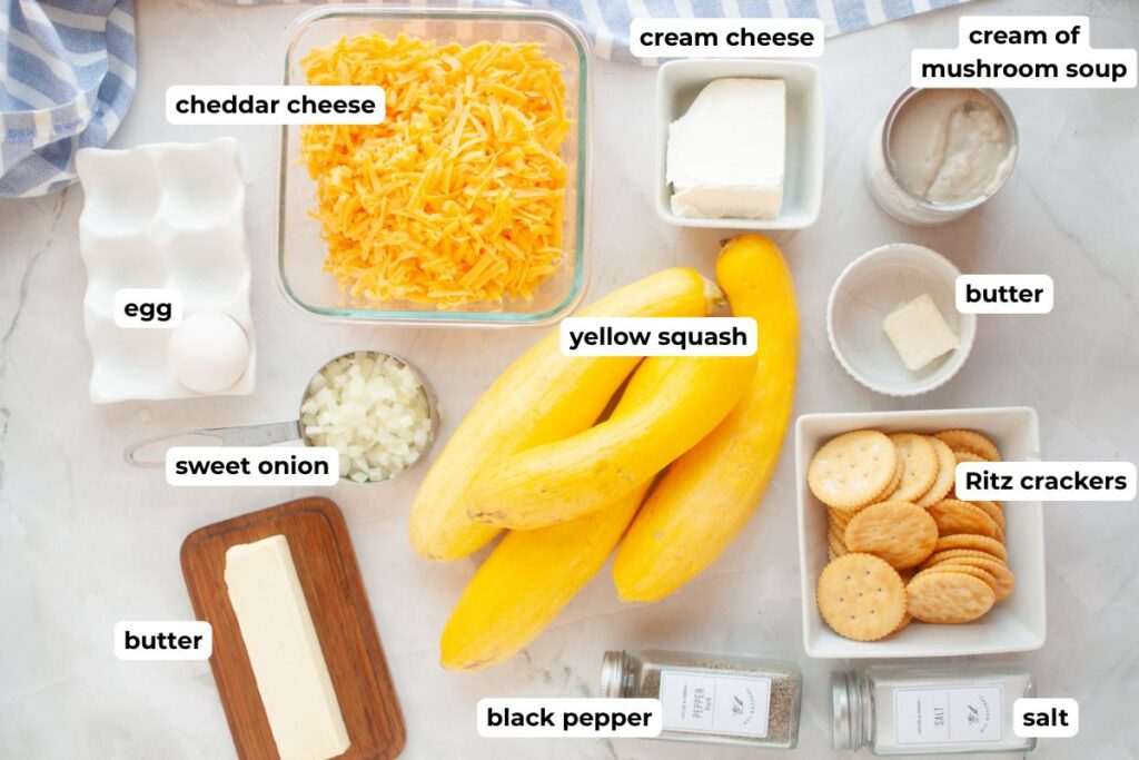 Top-down view of labeled ingredients for a recipe, including yellow squash, cheddar cheese, egg, sweet onion, butter, cream cheese, cream of mushroom soup, Ritz crackers, salt, and black pepper.