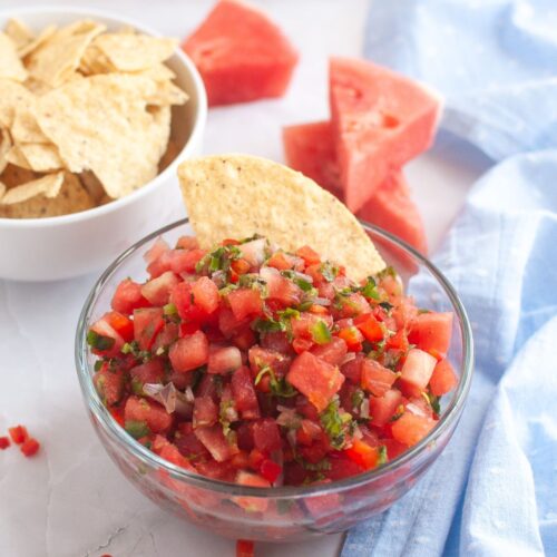 Fresh watermelon salsa in a glass bowl, garnished with chopped herbs and served with a tortilla chip, accompanied by a bowl of chips and watermelon slices in the background.