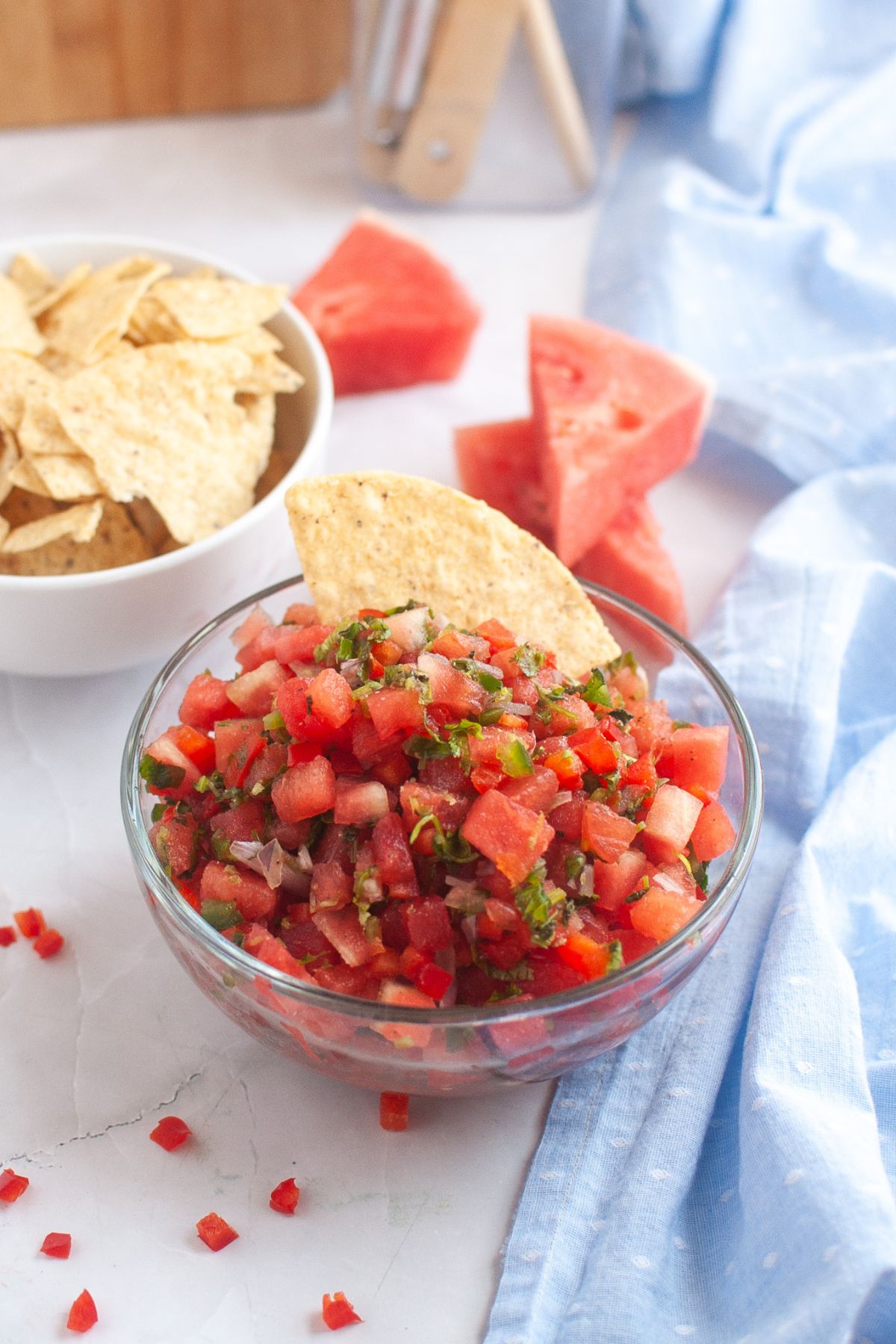 Fresh watermelon salsa in a glass bowl, garnished with chopped herbs and served with a tortilla chip, accompanied by a bowl of chips and watermelon slices in the background.