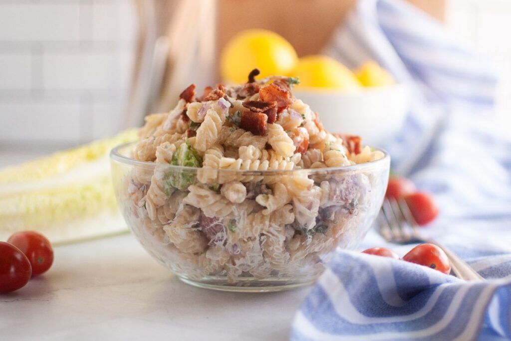 A glass bowl filled with BLT pasta salad, featuring spiral pasta, creamy dressing, pieces of bacon, and other visible ingredients, is shown on a light surface. Red onions, lettuce, cherry tomatoes, and a blue striped cloth are visible in the background.