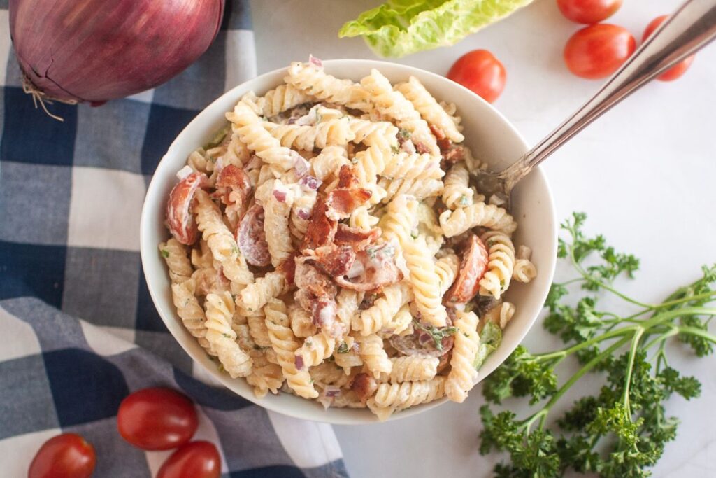 A white bowl filled with pasta salad, featuring spiral pasta, creamy dressing, pieces of bacon, and other visible ingredients, is shown with a spoon. Red onions, lettuce, cherry tomatoes, and parsley are in the background.