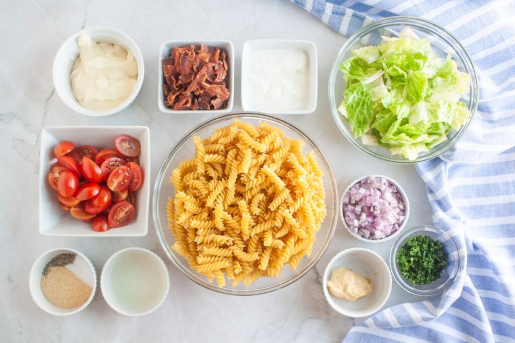 Overhead shot of various ingredients for a pasta salad, including cooked spiral pasta in a glass bowl, bowls of bacon, lettuce, red onion, parsley, tomatoes, and dressing components, arranged on a marble surface.