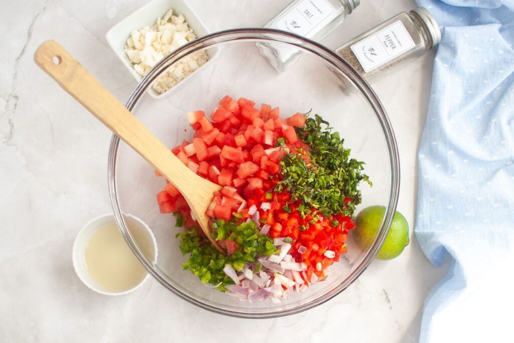 Overhead shot of a glass bowl containing diced watermelon, red bell pepper, shallots, jalapeno, and cilantro, with a wooden spoon inside. Other ingredients, including feta cheese, lime juice, lime, salt, and pepper, are visible around the bowl.