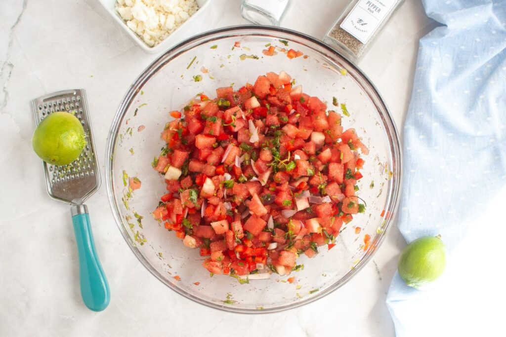 Overhead shot of a glass bowl filled with watermelon salad, featuring diced watermelon, red bell pepper, shallots, jalapeno, and cilantro. A grater and limes are visible on the side.