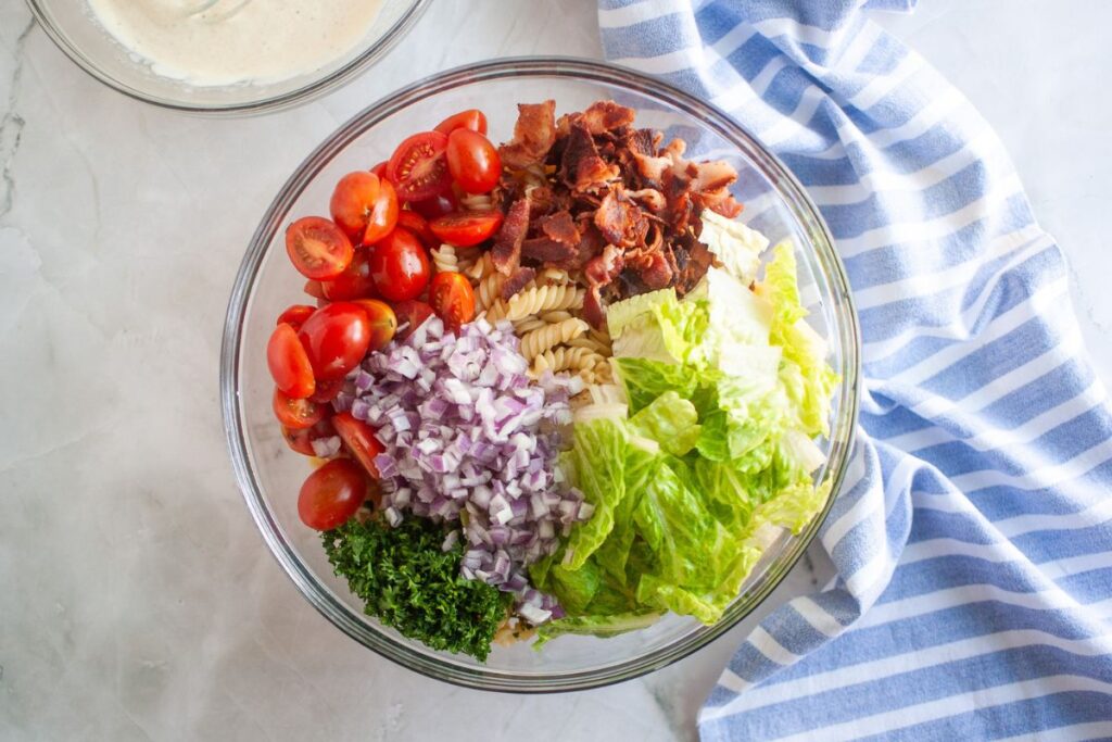 Overhead shot of a glass bowl containing BLT pasta salad ingredients, including pasta, tomatoes, bacon, lettuce, red onion, and parsley. A bowl of dressing and a blue striped cloth are also visible.