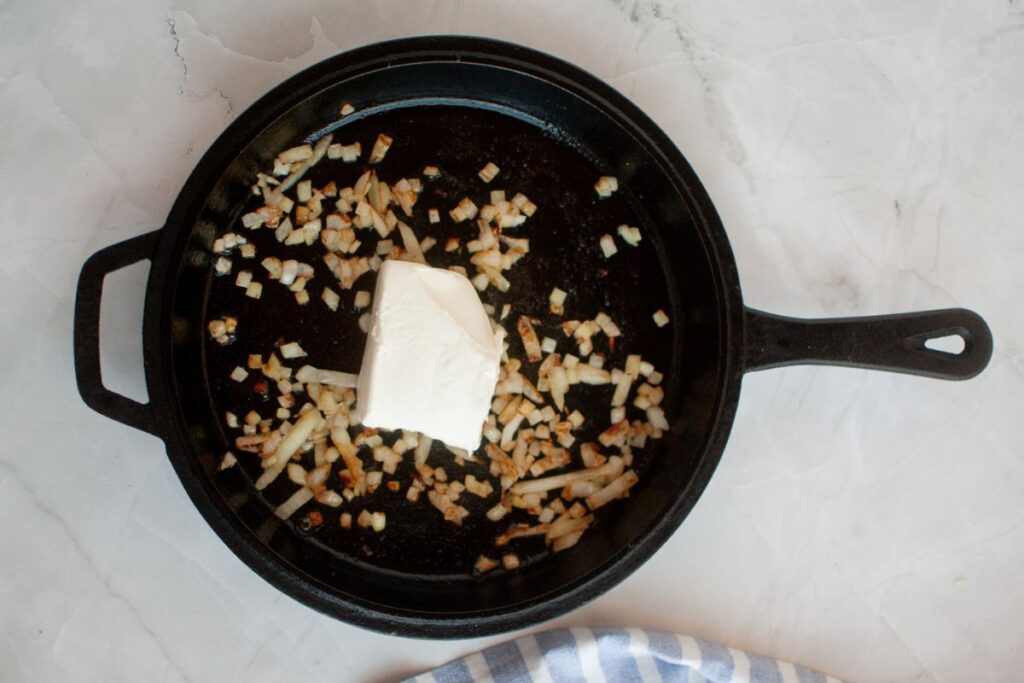 Chopped onions and a block of cream cheese in a black cast iron skillet on a light countertop.