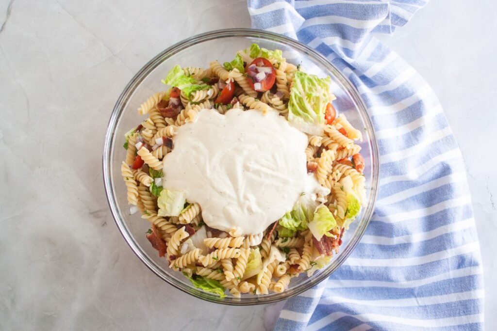 Overhead shot of a glass bowl filled with pasta salad, featuring spiral pasta, tomatoes, bacon, lettuce, and red onion, with a dollop of creamy dressing on top. A blue striped cloth is visible on the side.