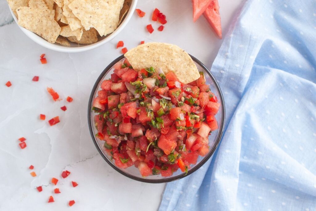 Overhead shot of a glass bowl filled with watermelon salsa, featuring diced watermelon, red bell pepper, shallots, jalapeno, and cilantro. A tortilla chip rests on top. A bowl of chips and a blue cloth are also visible.